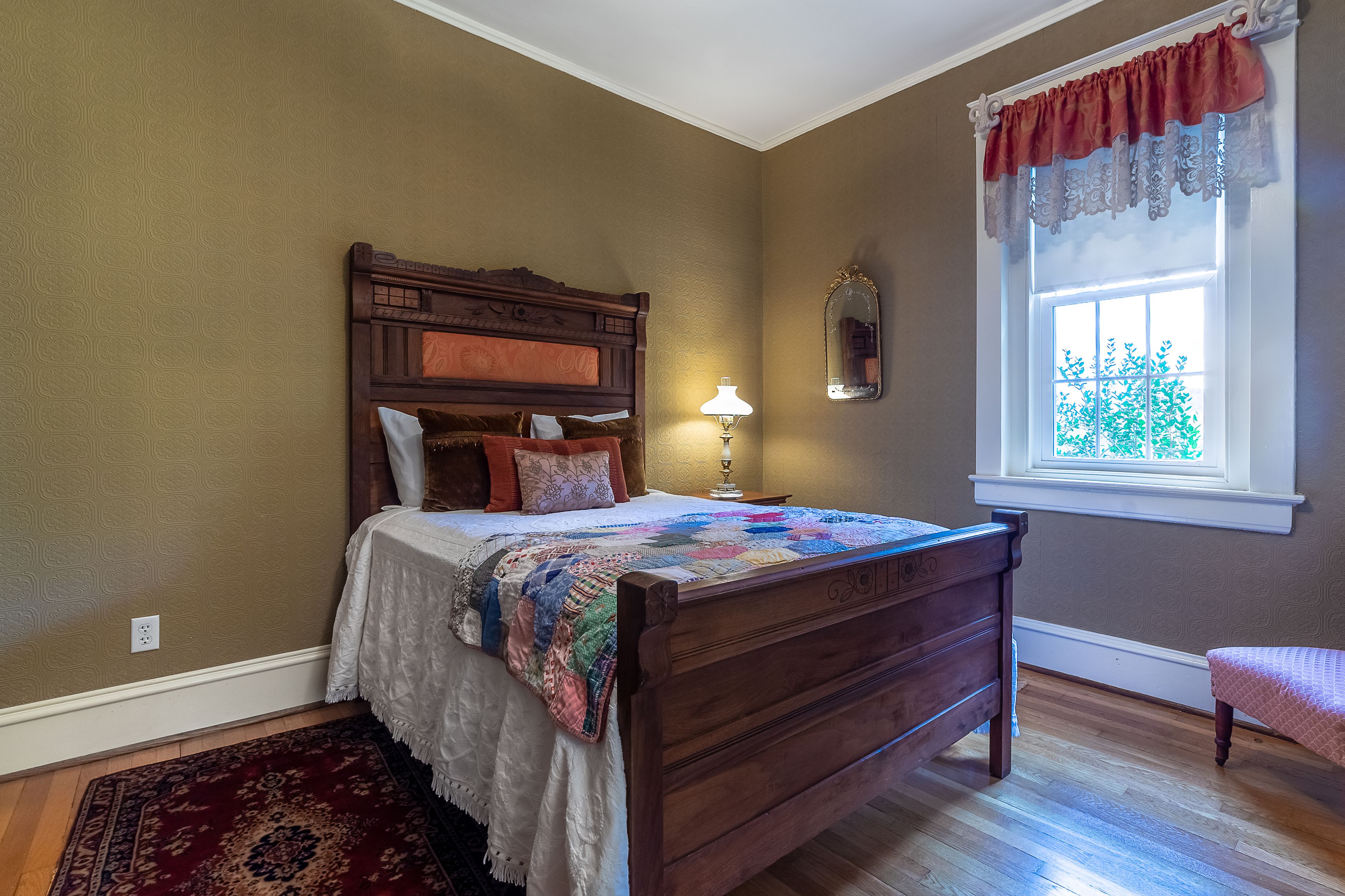 Une chambre avec un lit en bois, une table de chevet, une lampe, une fenêtre donnant sur un paysage verdoyant et un tapis à motifs.