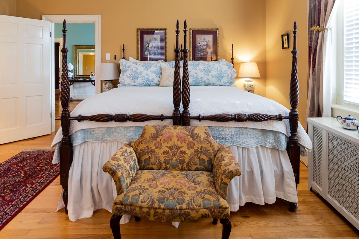 A four-poster bed with a floral-patterned chair in front, a red rug, and a window with curtains.