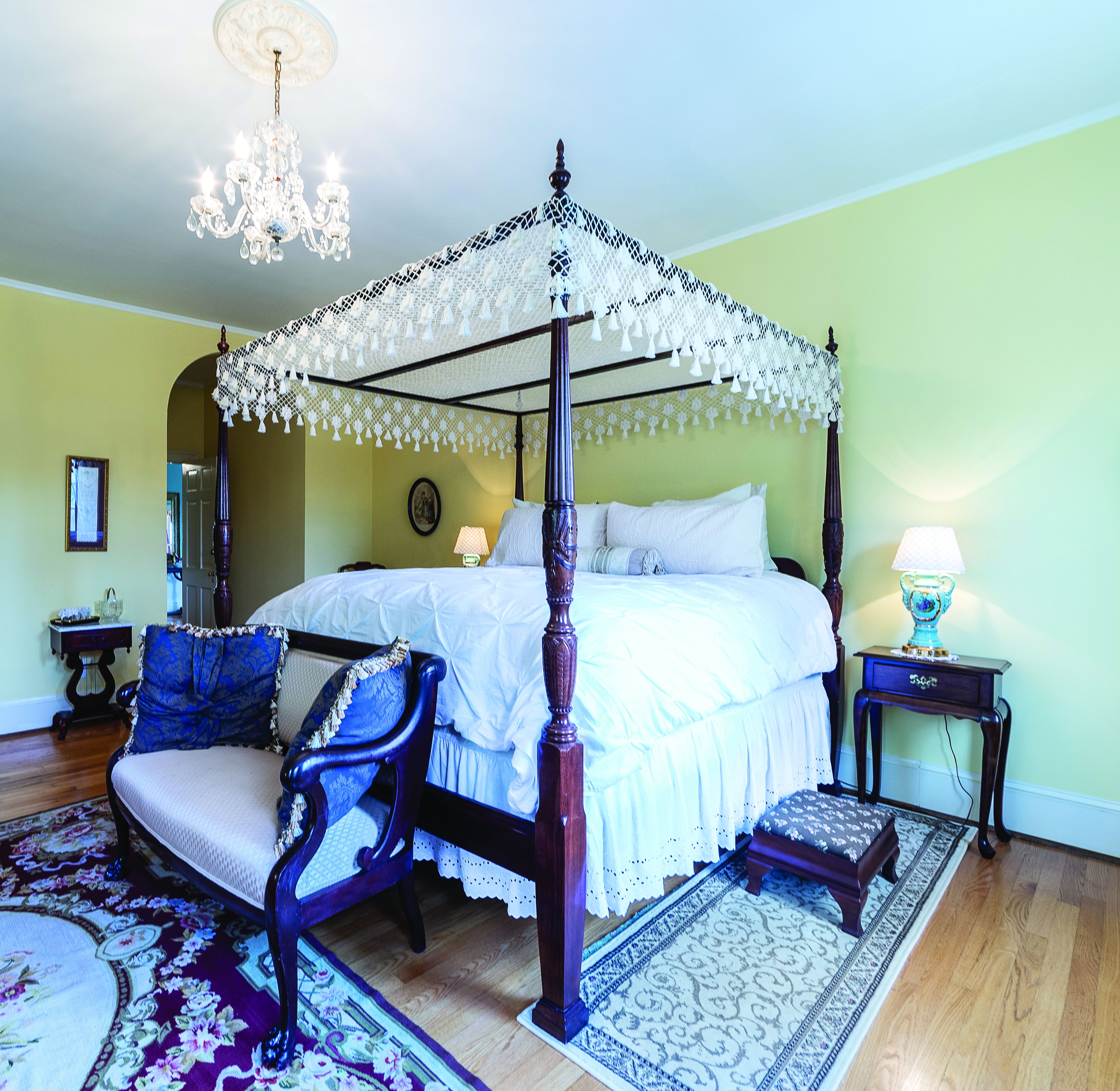 A four-poster bed with a canopy, a chair with a blue cushion, a bedside table with a lamp, and a patterned rug.