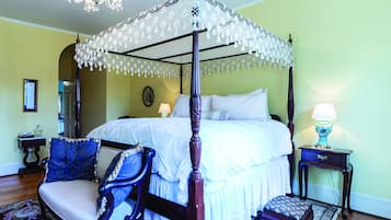 A four-poster bed with a canopy, a chair with a blue cushion, a bedside table with a lamp, and a patterned rug.