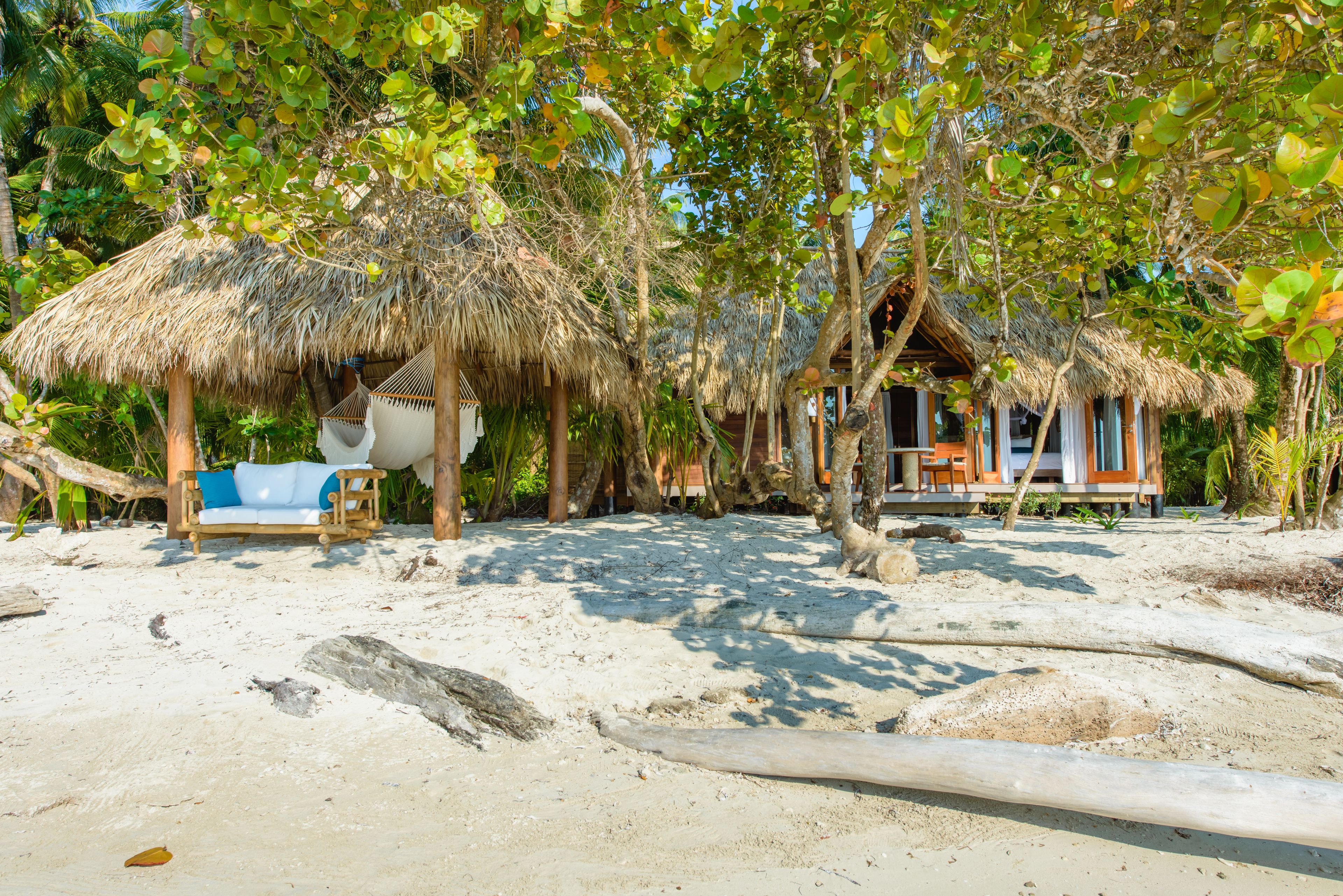 Un resort frente a la playa con cabañas de techo de paja, una banca de madera y una playa arenosa.
