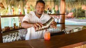 A bartender preparing a drink at a bar with a thatched roof, shelves with bottles, and a view of a pool in the background. - Calala Island (Laguna de Perlas)