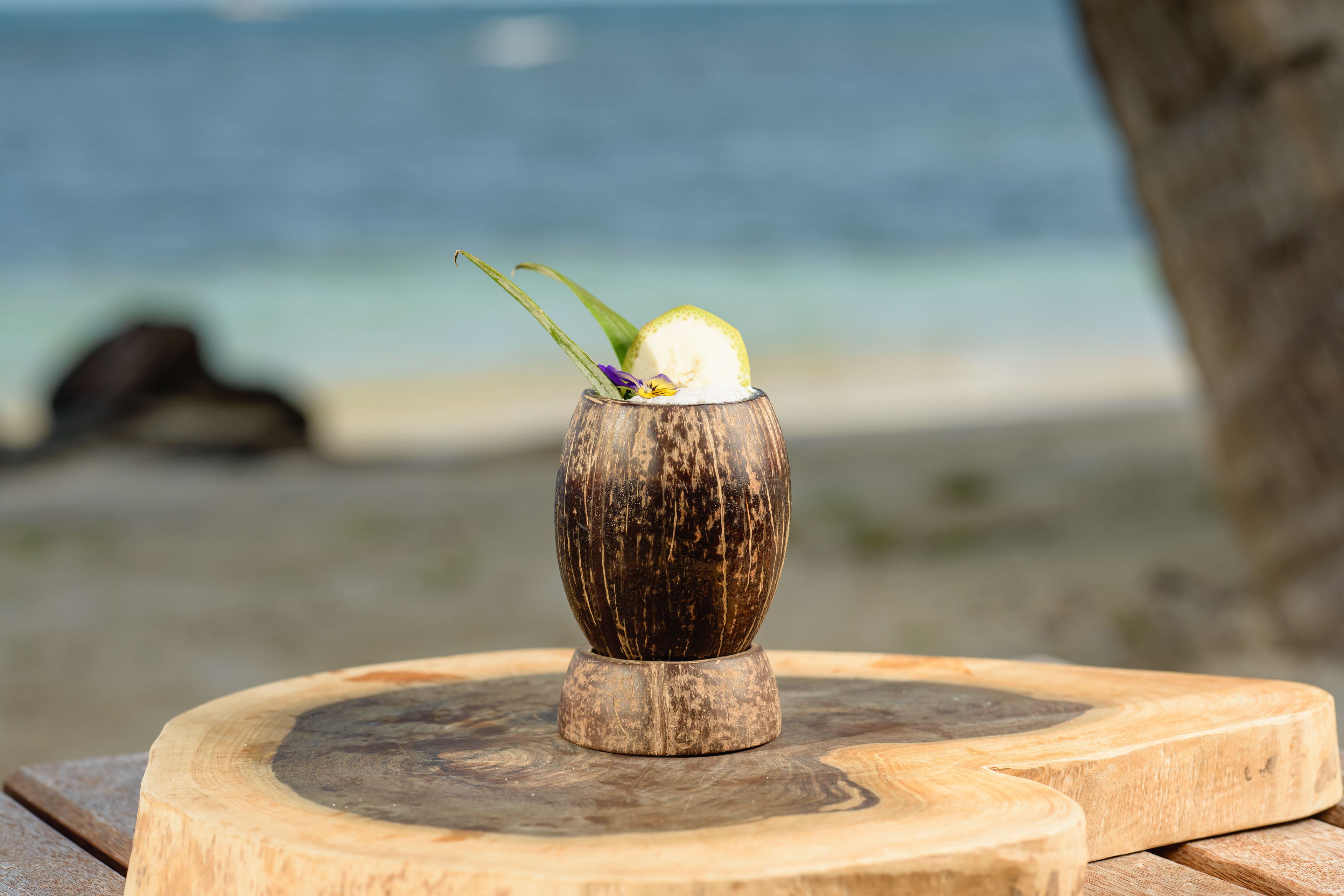 Una mesa de madera con un vaso que contiene una bebida adornada con una rodaja de limón y una flor, sobre una tabla de madera, con una playa y el océano al fondo.