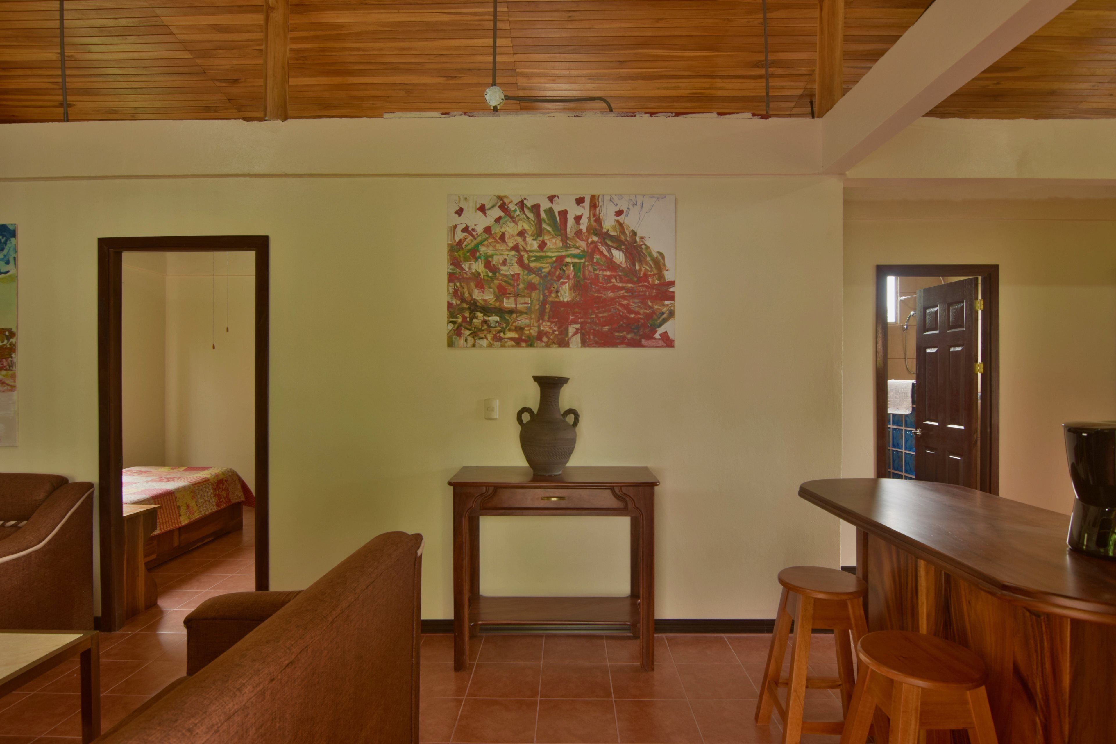 A room with a wooden ceiling, a bed with a red blanket, a wooden table with a vase, and a bar area with stools.