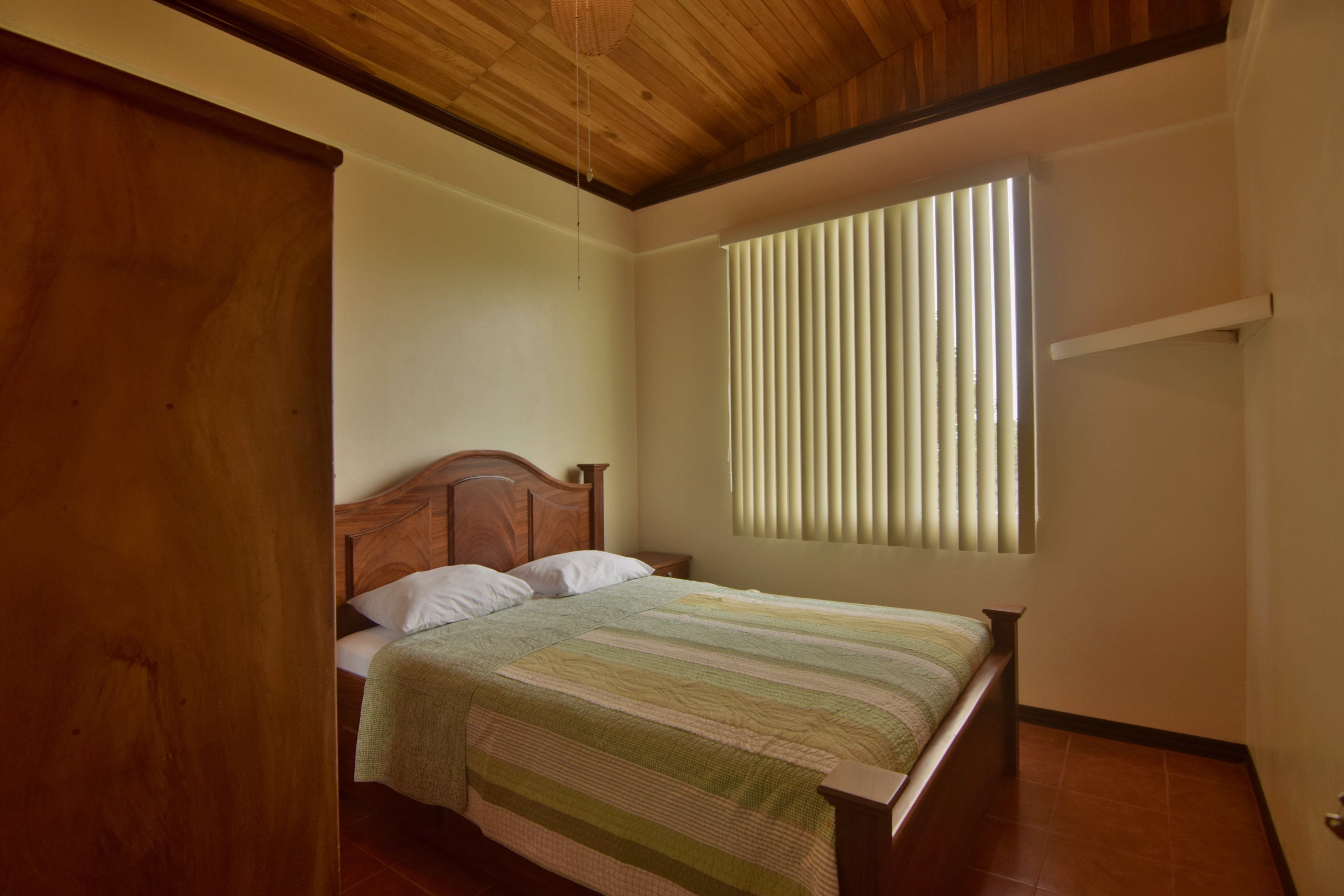 A bedroom with a wooden ceiling, a bed with a striped bedspread, a window with vertical blinds, and a small shelf above the bed.