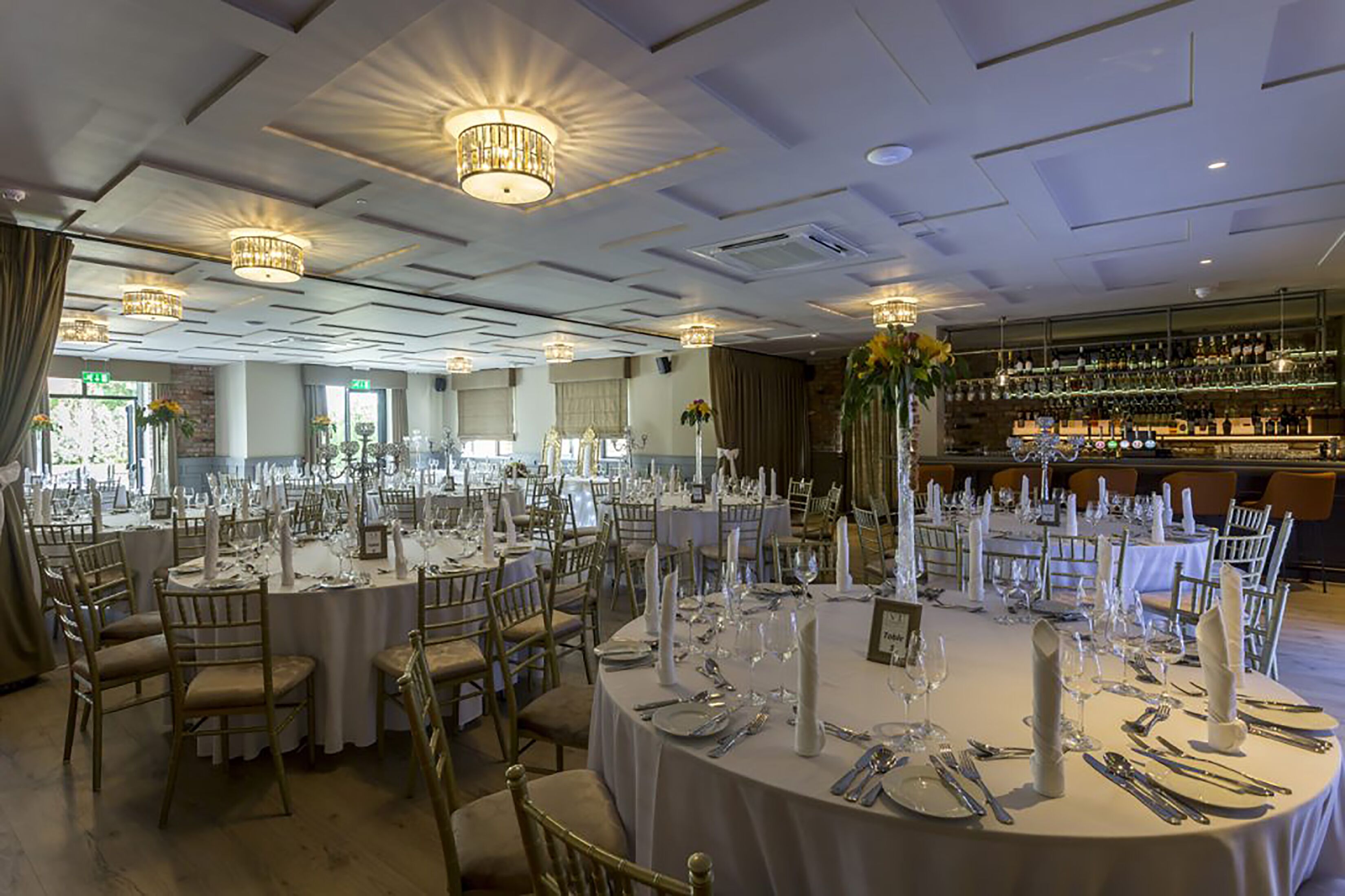 Una sala da banchetti con tavoli rotondi apparecchiati per un evento formale, con lampadari e un'area bar sullo sfondo.