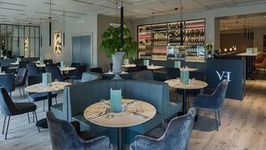 A modern restaurant with round tables, blue booths, and a bar area with a wine display. - Village Hotel (Bettystown)