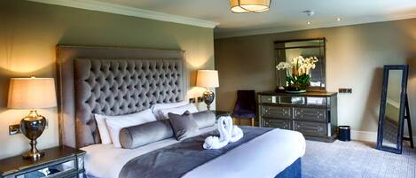 A hotel room with a tufted headboard, a large bed with white and gray bedding, two bedside lamps, a dresser with a mirror, and a blue patterned carpet.