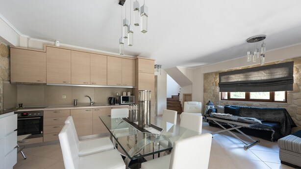 Une cuisine moderne avec une table à manger en verre et des chaises blanches, un four encastré et un mur d’accent en pierre.