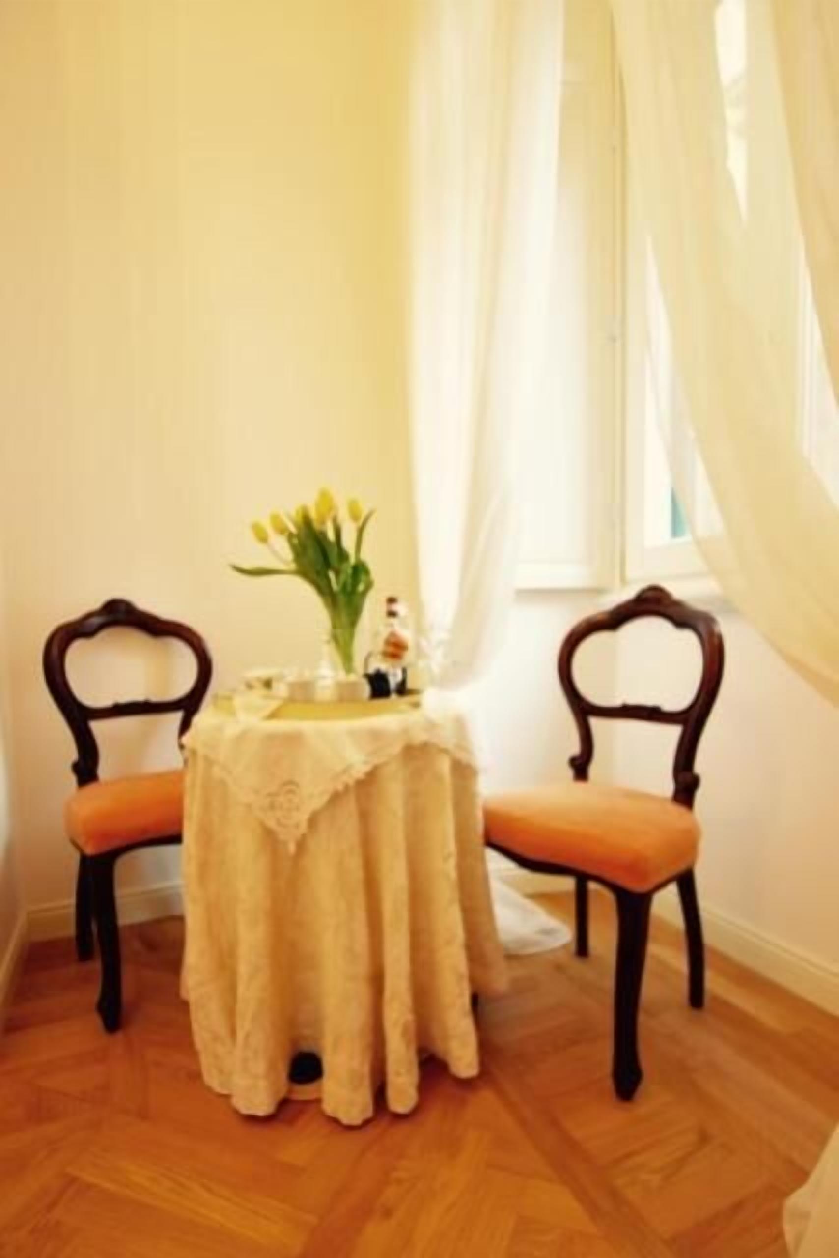 A room with two wooden chairs, a small round table with a white tablecloth, a vase with yellow flowers, and sheer white curtains.