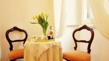 A room with two wooden chairs, a small round table with a white tablecloth, a vase with yellow flowers, and sheer white curtains.
