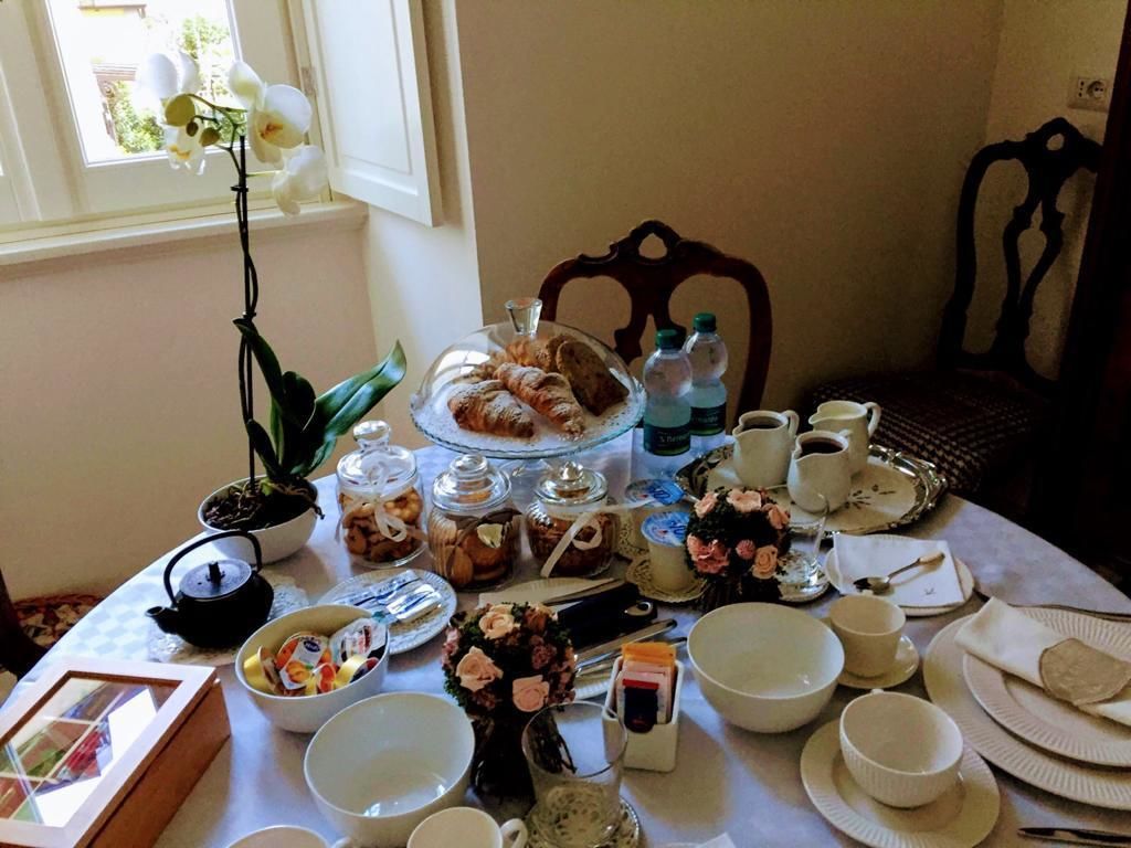A breakfast table set with various food items, including pastries, fruits, and beverages, in a room with a window and a chair.