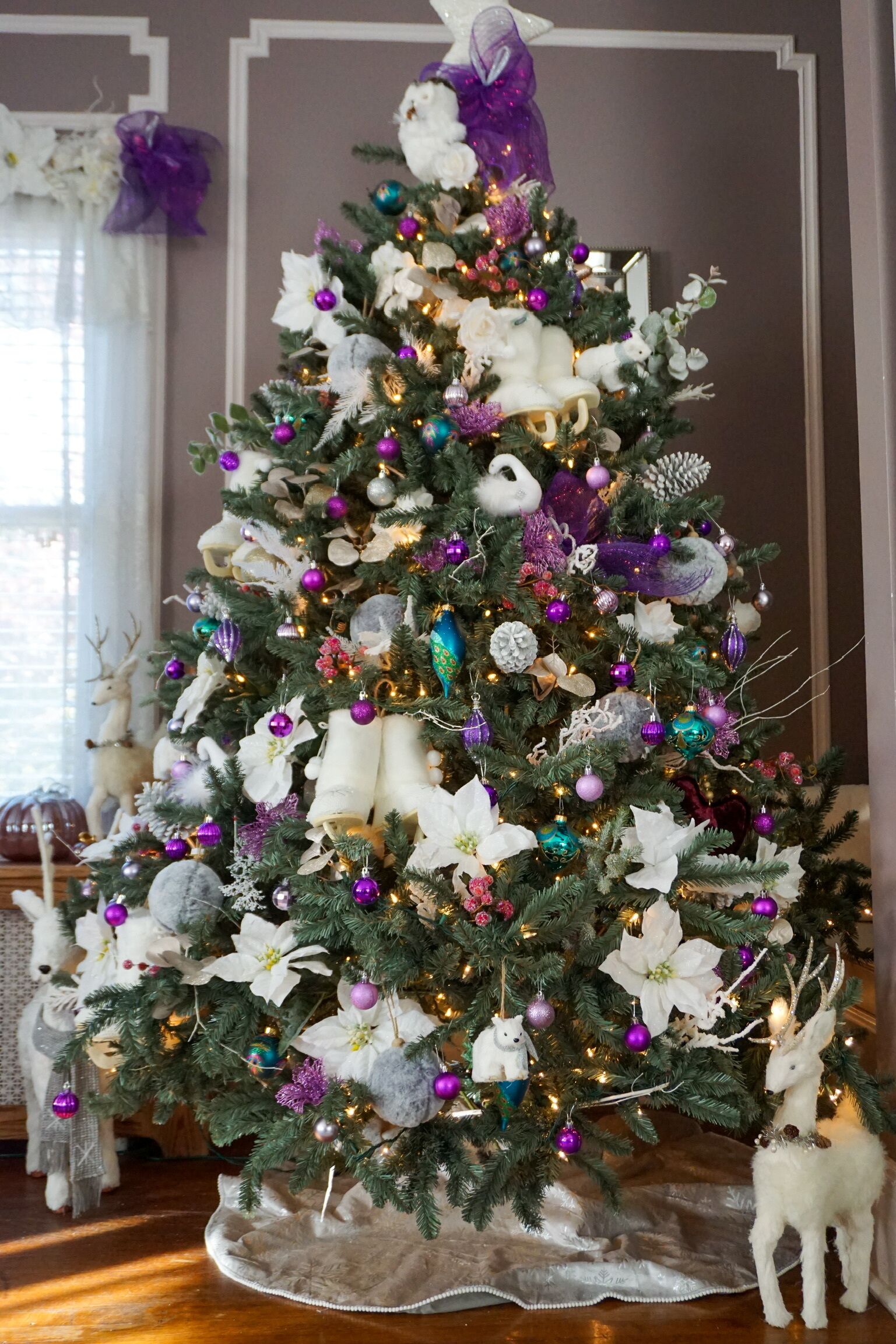 Een kerstboom met witte en paarse decoraties, omringd door een witte hertenbeeldje en een witte rendierbeeldje.