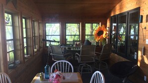 L’intérieur d’un chalet en bois, avec une table à manger, des chaises, une grande fenêtre ornée d’un tournesol et une vue sur les arbres.
