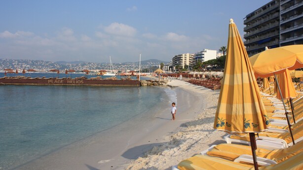 Une plage avec des transats et des parasols, un enfant qui marche le long du rivage, et un bâtiment en arrière-plan.