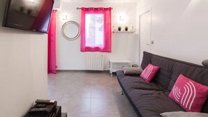 Ett vardagsrum med en soffa, en platt-TV som är monterad på väggen, ett element och ett fönster med en rosa gardin.