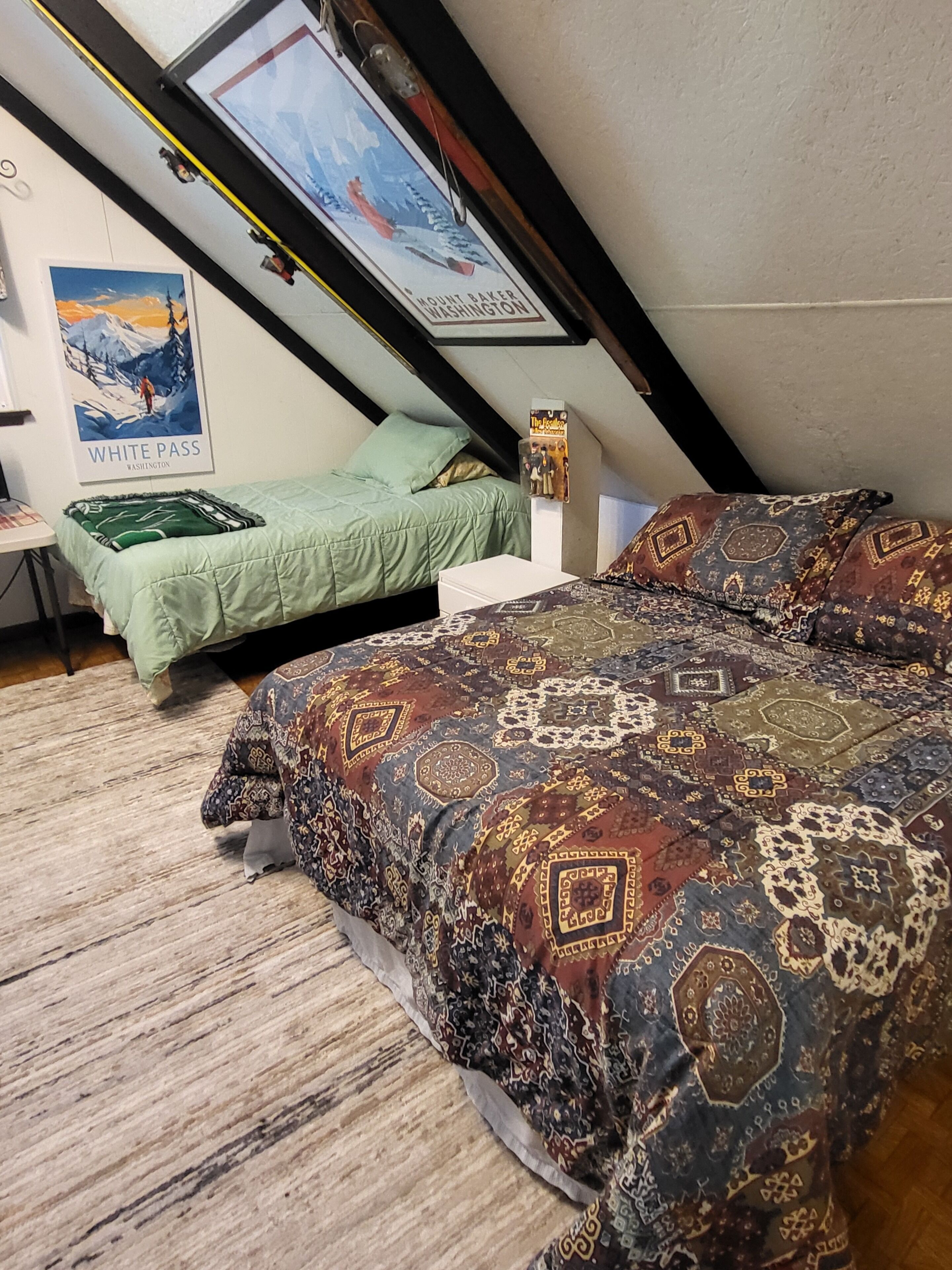 A loft bedroom with a sloped ceiling, a bed with patterned bedding, a green sofa, a small table, and a poster of Mount Baker, Washington.