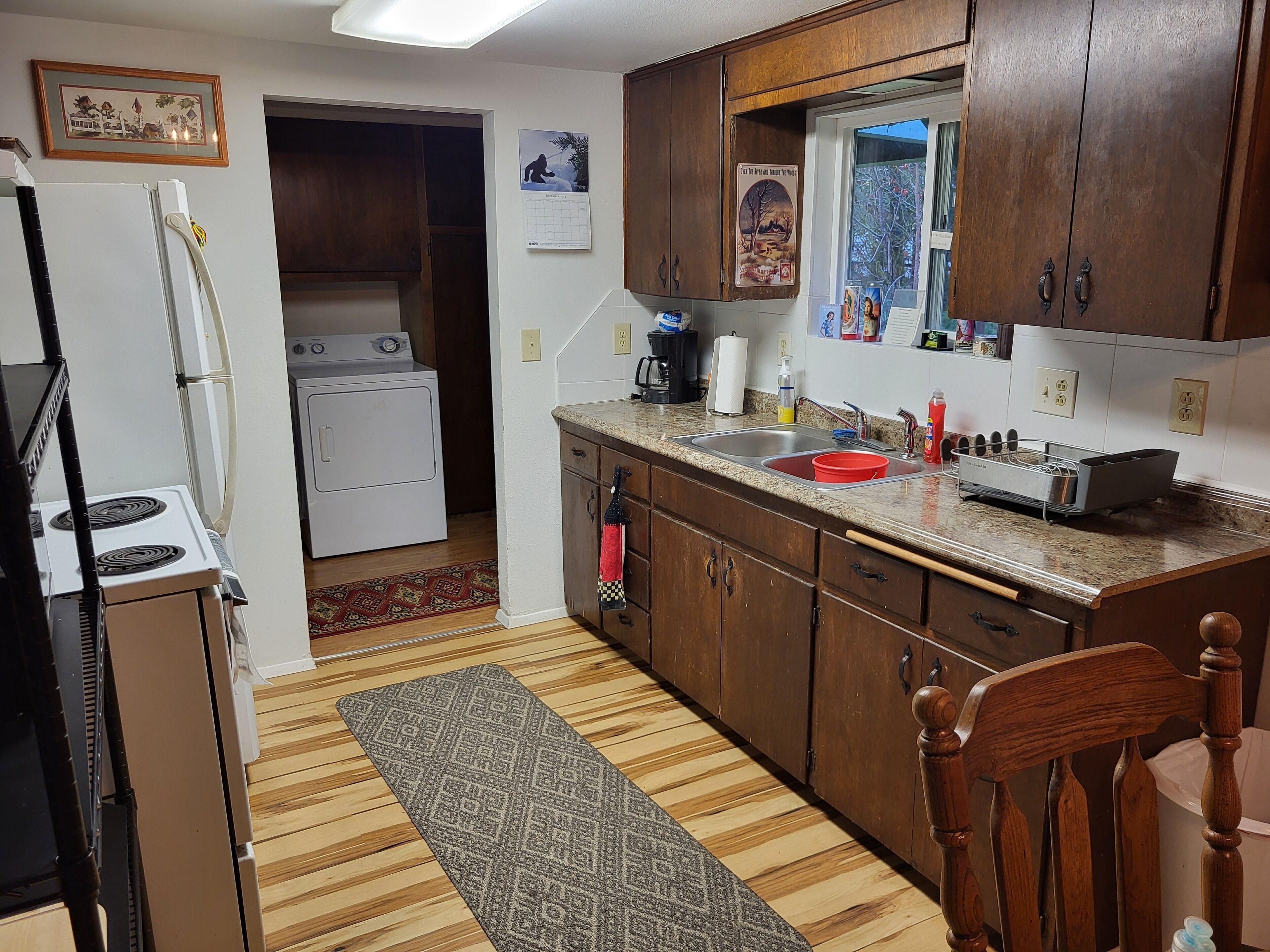 A compact kitchen with a stove, refrigerator, and sink. There is a wooden cabinet and a rug on the floor.