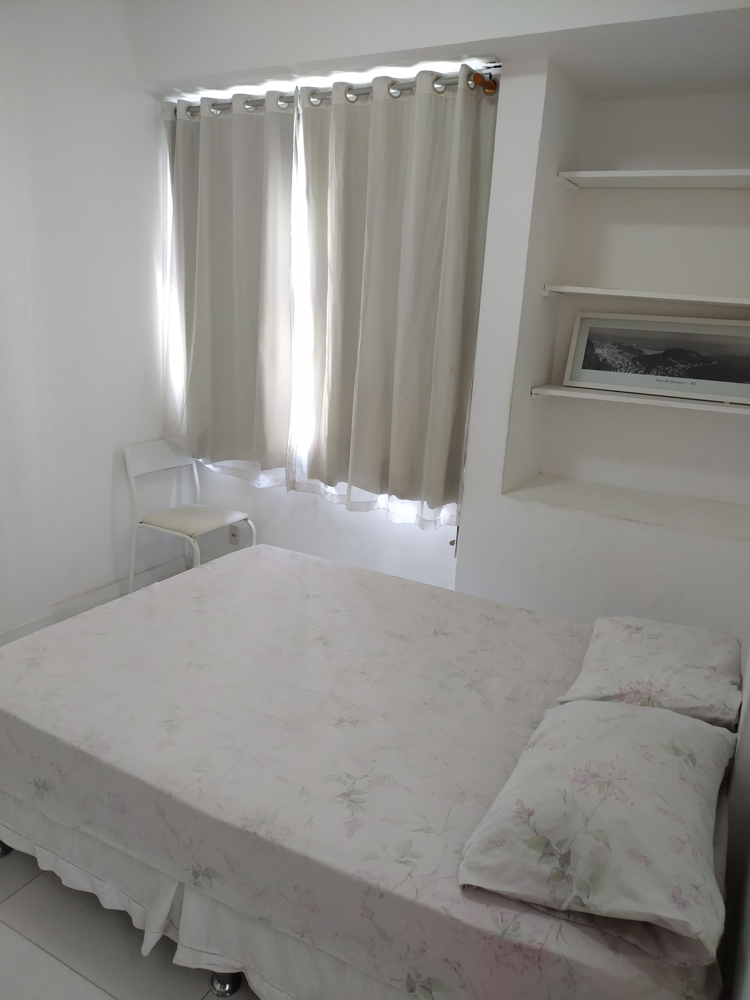 A bedroom with a bed, a chair, a shelf with a framed picture, and a window with curtains.