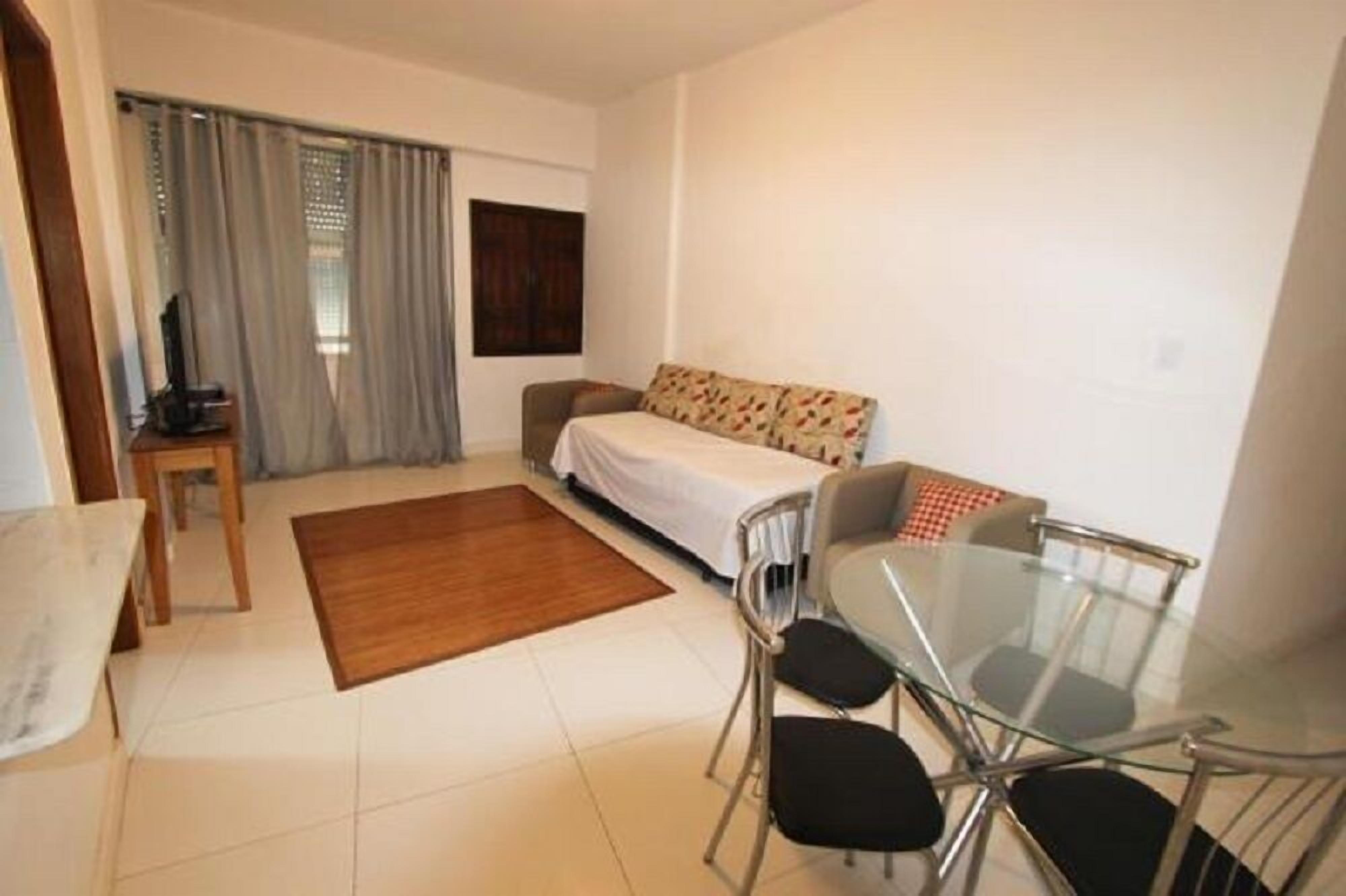 Een compacte, goed verlichte kamer met een bed, een slaapbank, een glazen eettafel met stoelen, een bijzettafeltje met een stoel, en een televisie op een standaard.