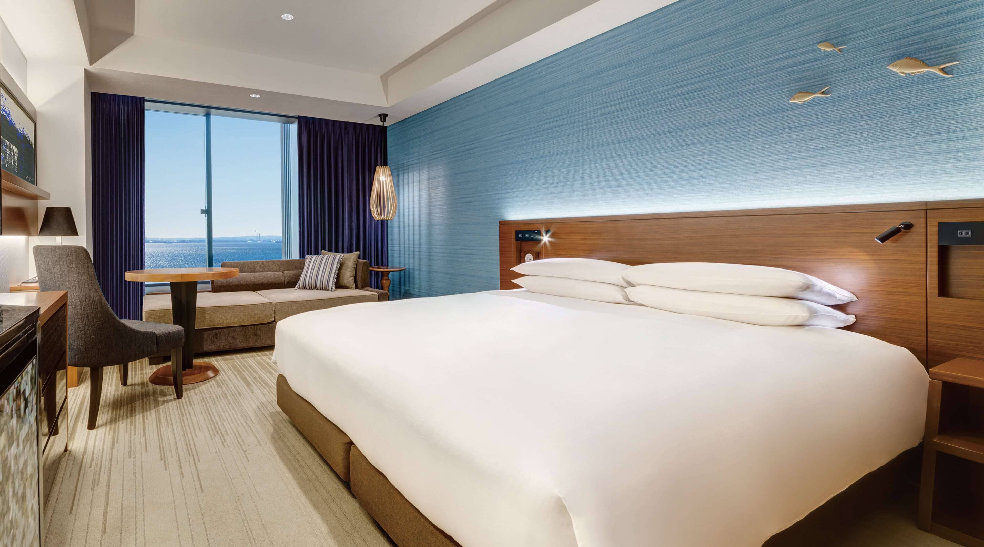 A hotel room with a large bed, a desk, a chair, a small table, and a view of the sea.