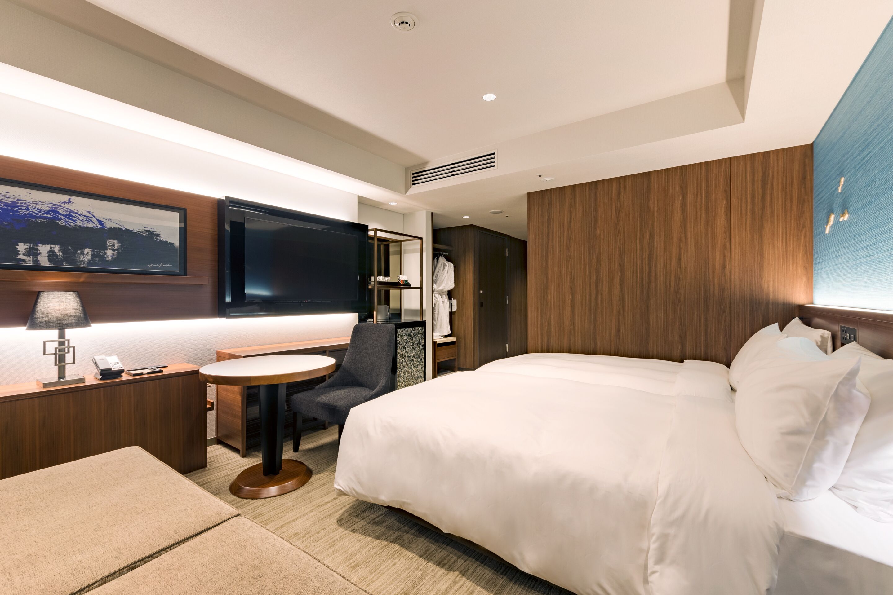 A modern hotel room with a large bed, a desk with a lamp, a chair, and a television.