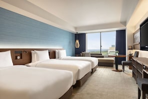 A hotel room with three beds, a desk, a chair, a TV, and a large window with a view.