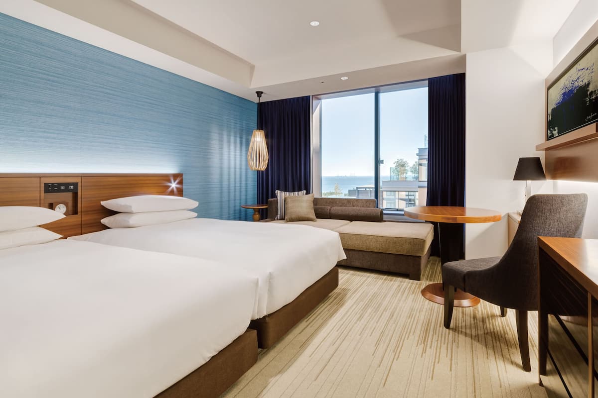 Ein Hotelzimmer mit zwei Betten, einem Schlafsofa, einem Sessel, einem kleinen Tisch und Blick auf die Stadt.
