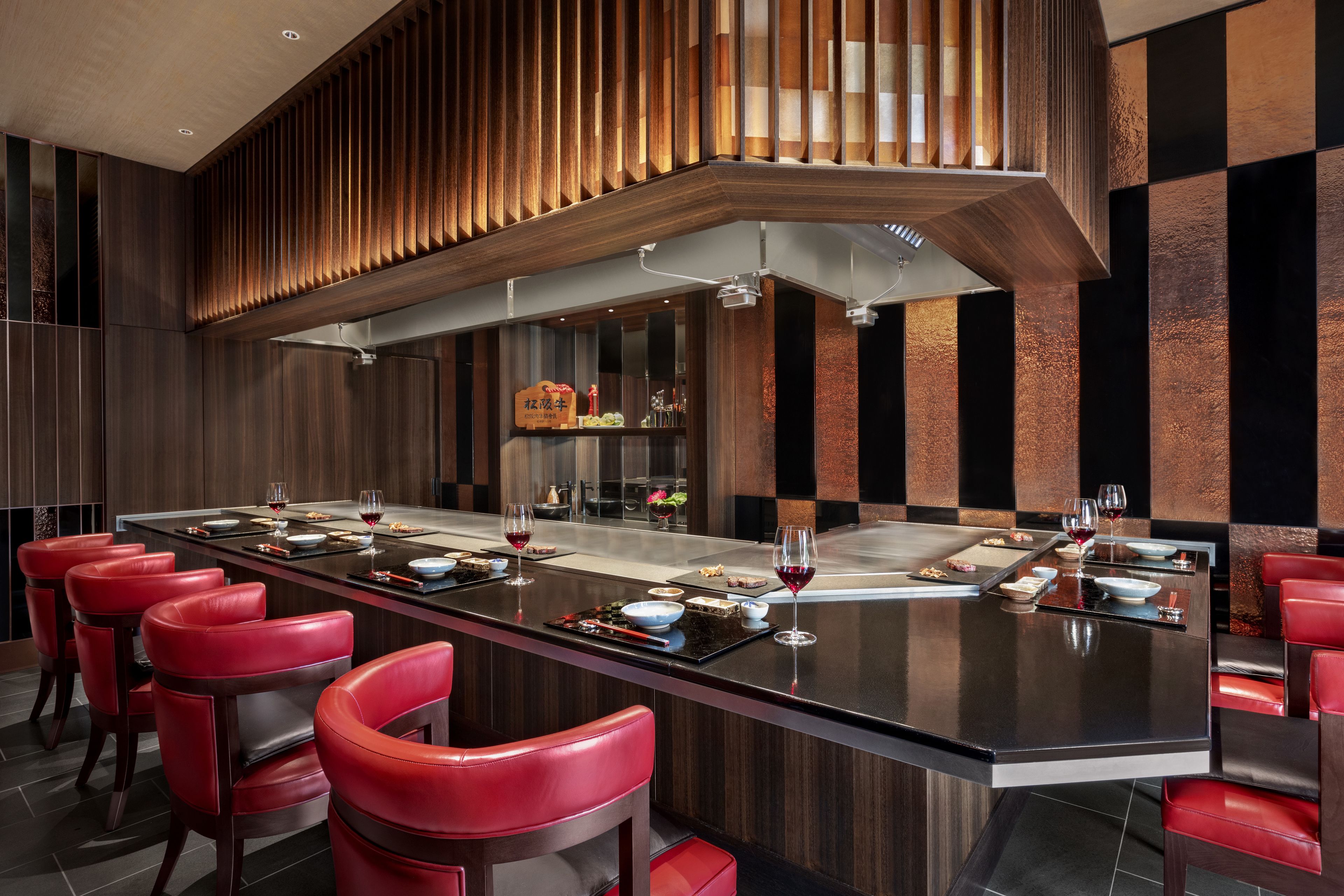A modern restaurant with a long dining table, red chairs, and a high ceiling with wooden slats.