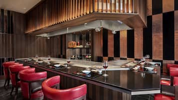A modern restaurant with a long dining table, red chairs, and a high ceiling with wooden slats.