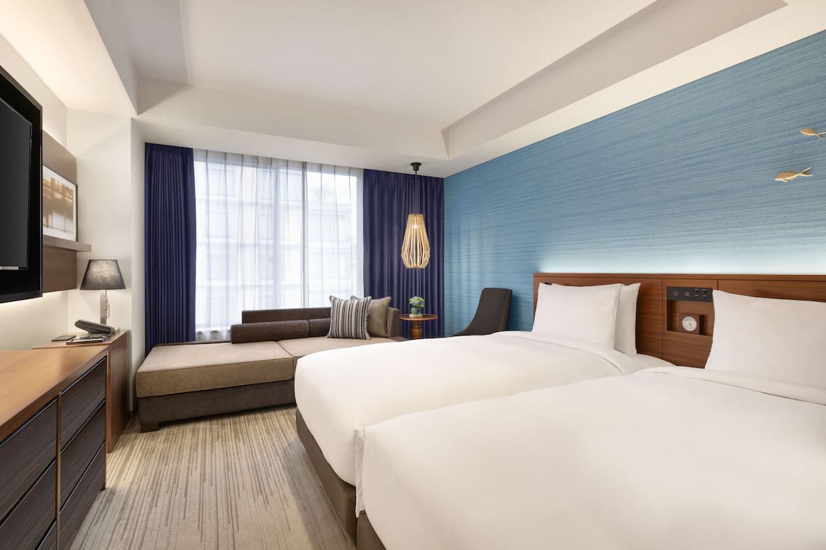 Ein Hotelzimmer mit zwei Betten, einem Schreibtisch, einem Fernseher und einem Fenster mit Vorhängen.