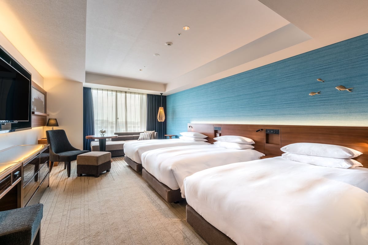 Ein Hotelzimmer mit drei Betten, einem Schreibtisch, einem Fernseher und einem Fenster mit Vorhängen.