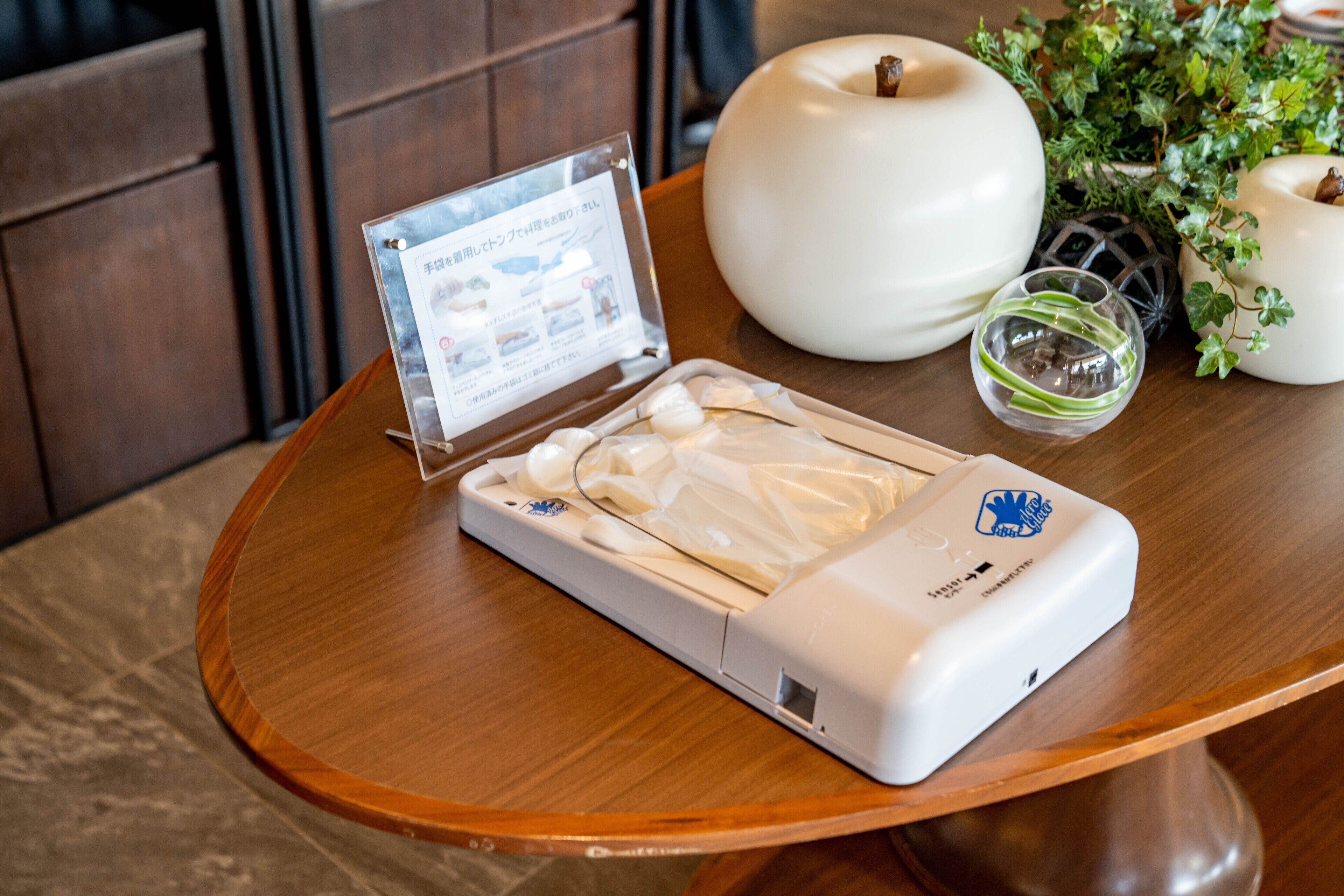 A table with a white device, a clear plastic sign, a white container, a glass, and a potted plant.