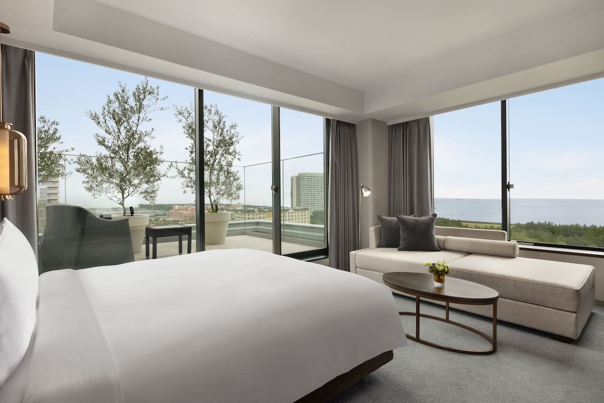 Ein modernes Hotelzimmer mit einem großen Bett, einer Sitzecke, einem Couchtisch und Blick auf Stadt und Meer.