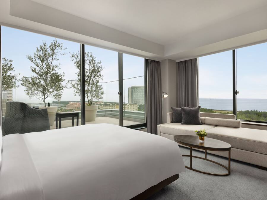 A modern hotel room with a large bed, a sofa, a coffee table, and a view of the city and sea.