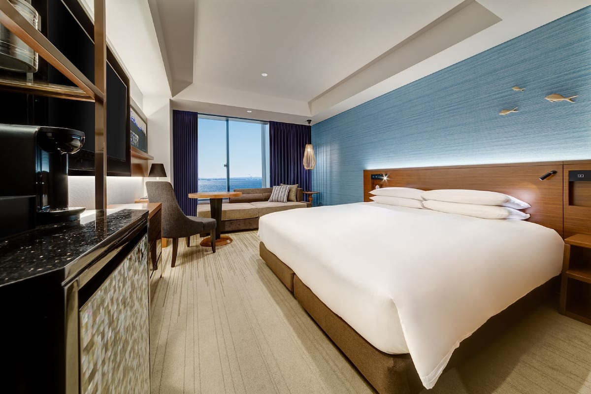 Ein modernes Hotelzimmer mit einem großen Bett, einem Schreibtisch, einem Stuhl, einer Kaffeemaschine und Blick auf die Stadt.