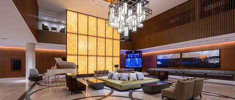 A spacious lobby with a modern chandelier, comfortable seating, a large TV screen, and a high ceiling with wooden paneling.