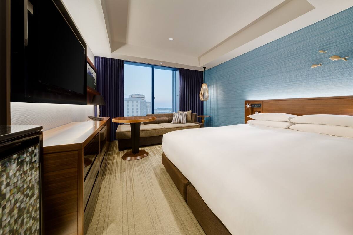 Ein Hotelzimmer mit einem großen Bett, einem Schreibtisch, einem Fernseher und Blick auf die Stadt durch große Fenster.