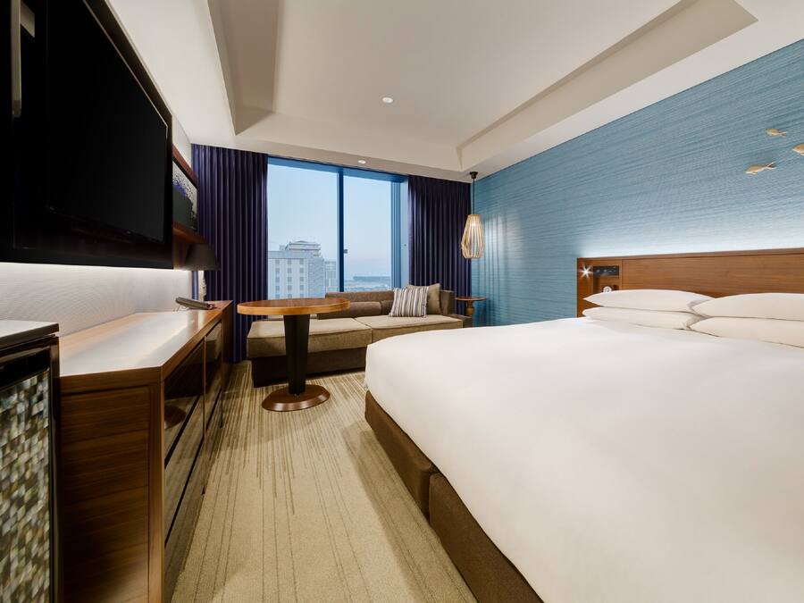 Hotelkamer met een groot bed, een bureau, een televisie en uitzicht op de stad door grote ramen.