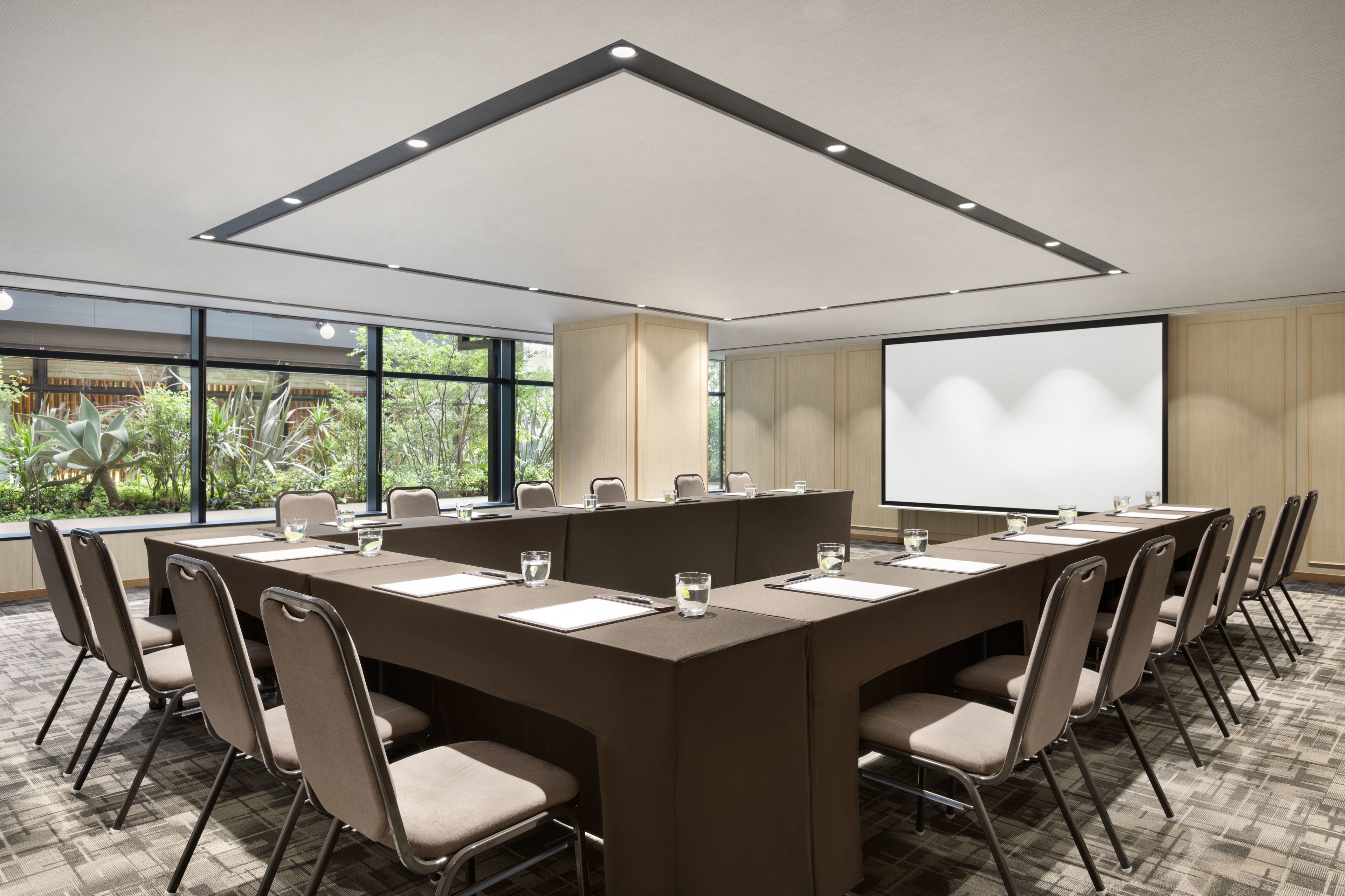 Un salón de conferencias con una larga mesa, sillas, una pantalla de proyector y un amplio ventanal con vistas al verdor.
