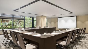 A conference room with a long table, chairs, a projector screen, and a large window with a view of greenery.