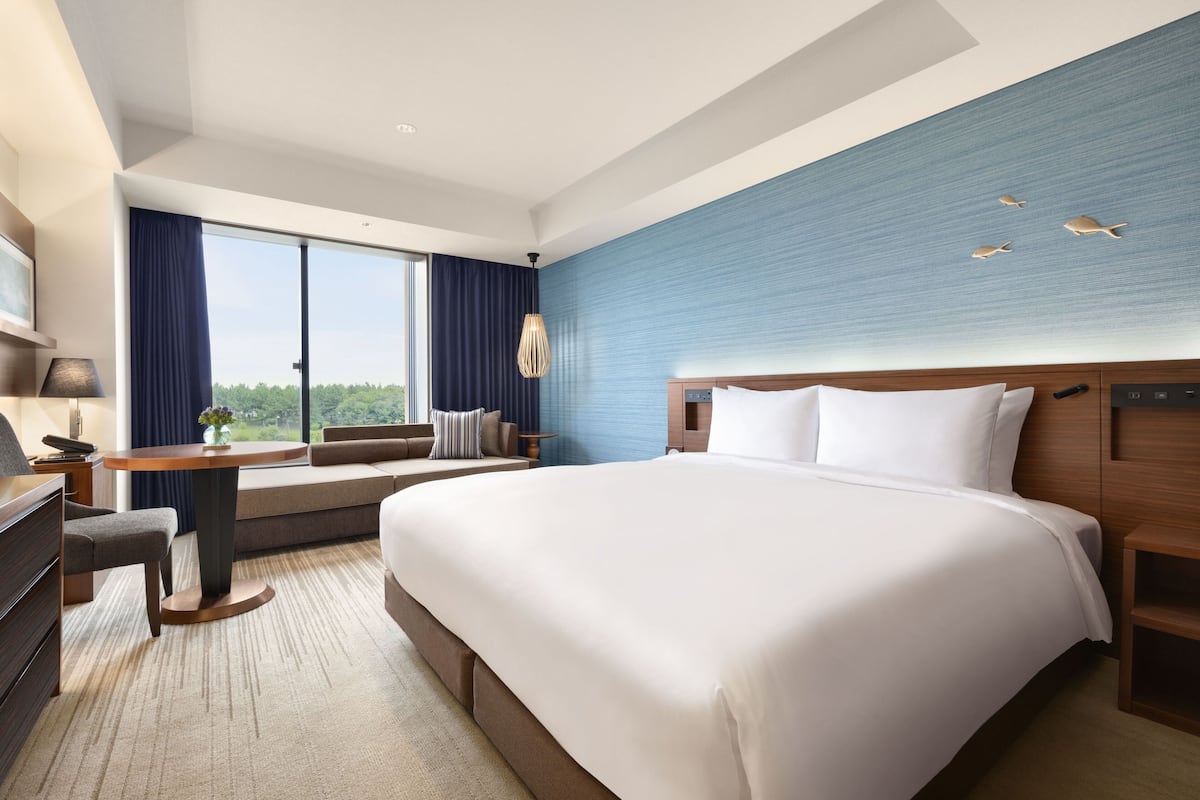 Ein modernes Hotelzimmer mit einem großen Bett, einem Schreibtisch mit Stuhl, einer Couch und einem großen Fenster mit Blick ins Freie.