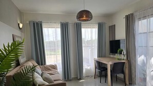 A modern living room with a sofa, a dining table with chairs, a TV mounted on the wall, and large windows with curtains.