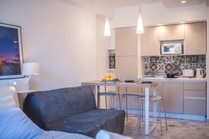Un salon moderne avec un canapé gris, une table à manger avec deux tabourets de bar et un coin cuisine équipé d’un four à micro-ondes et doté d’un dosseret carrelé.