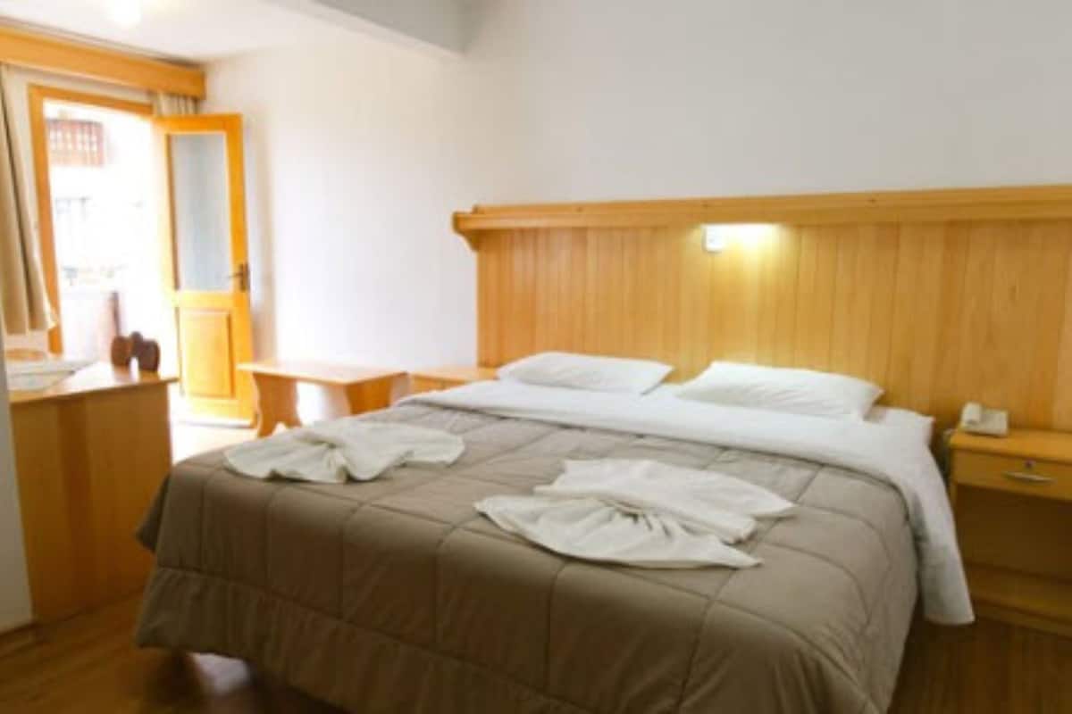 Une chambre d’hôtel avec une tête de lit en bois, un lit recouvert d’une couette beige, une table de chevet avec une lampe et une commode en bois.