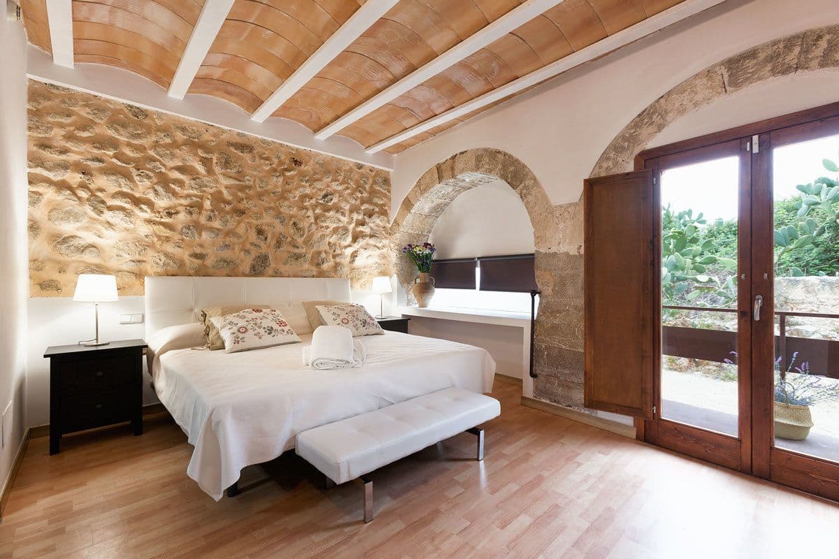 Un dormitorio con pared de piedra, cama con ropa de cama blanca, un banco, una mesita de noche y una ventana con vistas a zonas verdes.