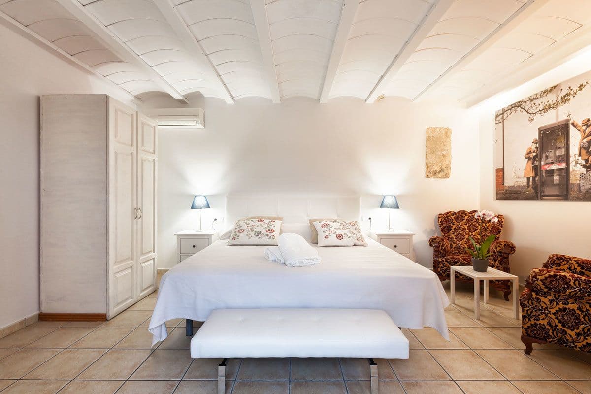 Un dormitorio con una cama grande, dos mesitas de noche con lámparas, un banco, un armario y un sillón estampado.