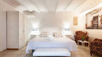 Un dormitorio con una cama grande, dos mesitas de noche con lámparas, un banco, un armario y un sillón estampado.