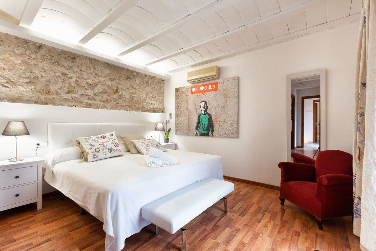 Un dormitorio con cama, mesitas de noche, un banco, un sillón rojo, una pared de piedra y un cuadro.
