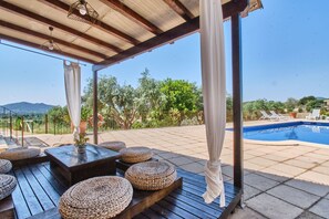 An outdoor patio area with wooden tables and woven ottomans, a pool, and a view of the landscape.